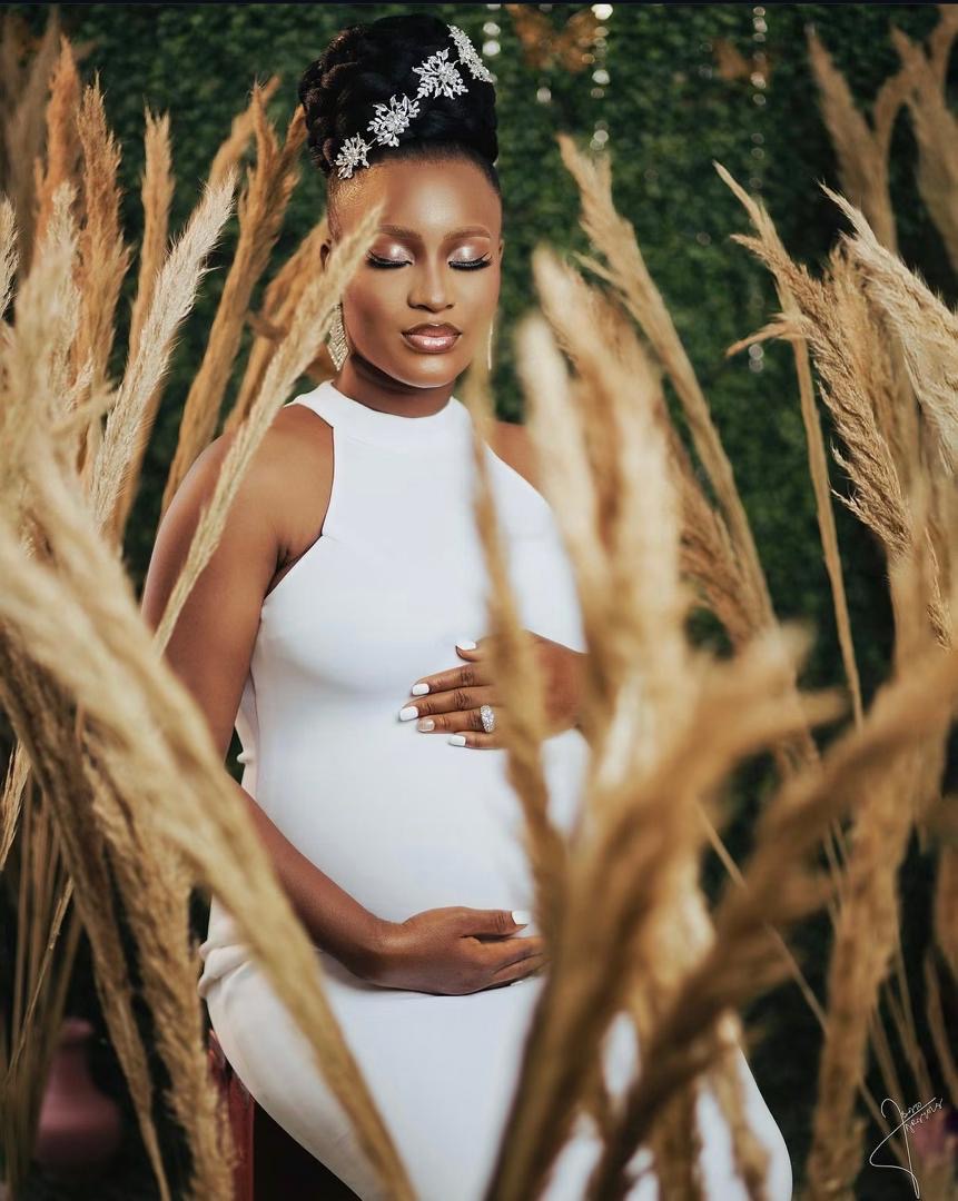 Expectant mother in a white dress gently cradling her bump among pampas grass