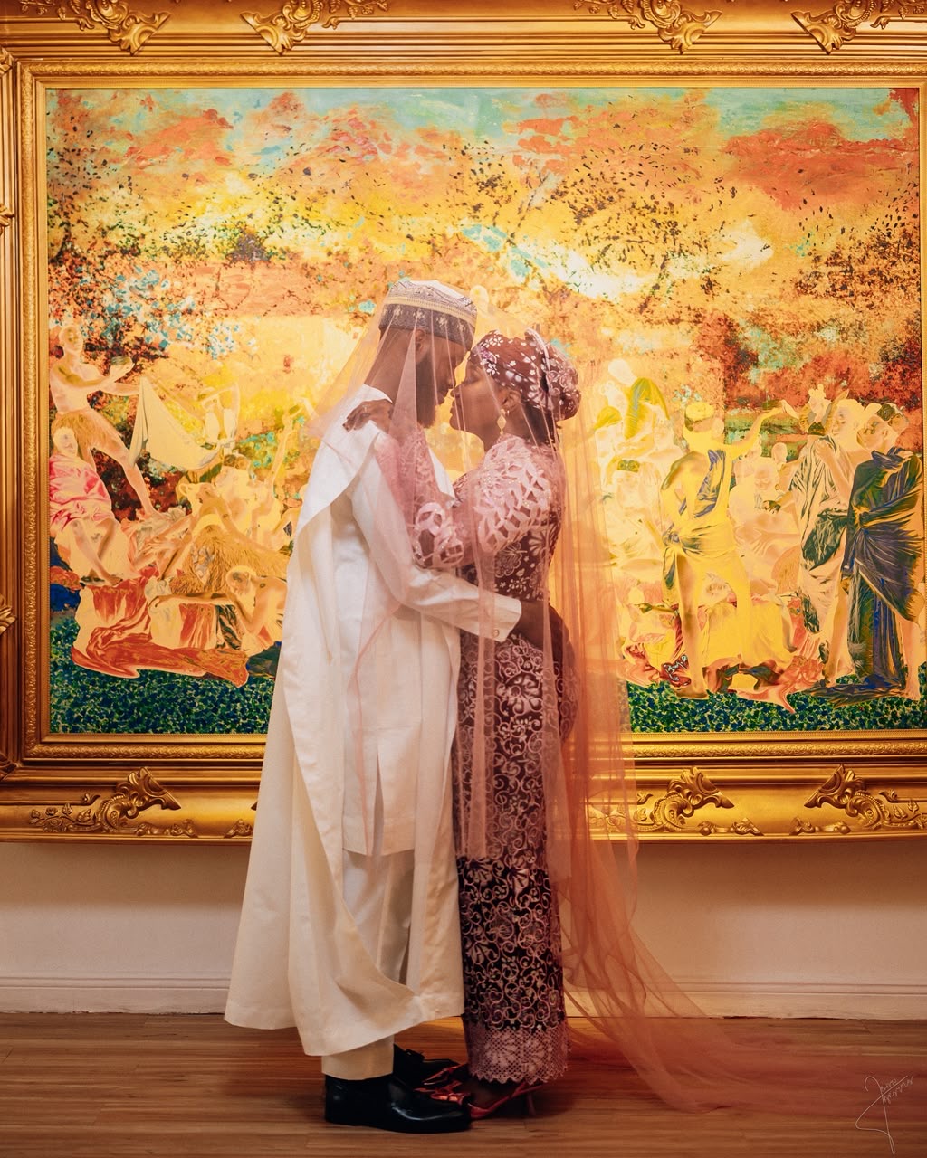 Couple embracing under a veil in traditional attire before a golden framed painting