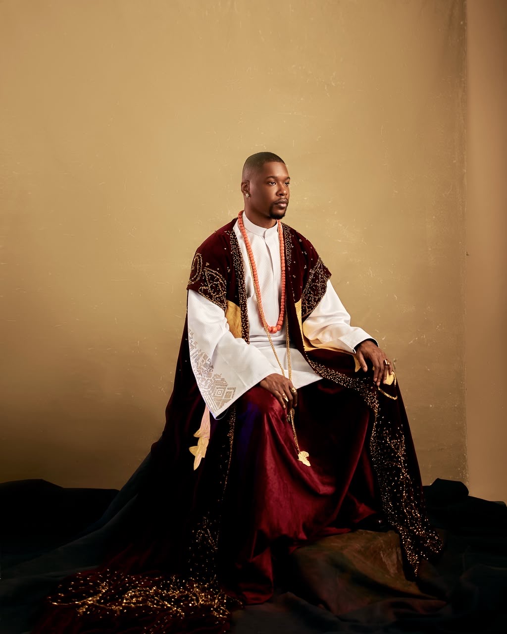 Seated portrait of a man in ornate burgundy and white attire with coral beads