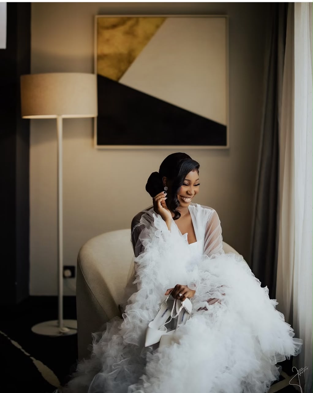 Smiling bride seated by a window in a white feathered robe